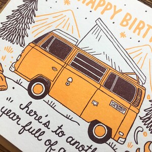 May include: A greeting card with a yellow vintage van with a pop-up roof, mountains, and trees in the background. The text on the card reads "Happy Birthday" and "Here's to another year full of..." with a sticker on the van that says "Wilderness or Bust!"