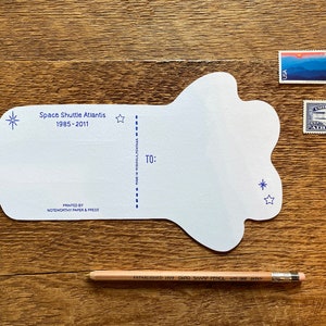 May include: A white postcard shaped like a cloud with blue stars and the text "Space Shuttle Atlantis 1985-2011" printed on it. The postcard has a space for a message and an address.