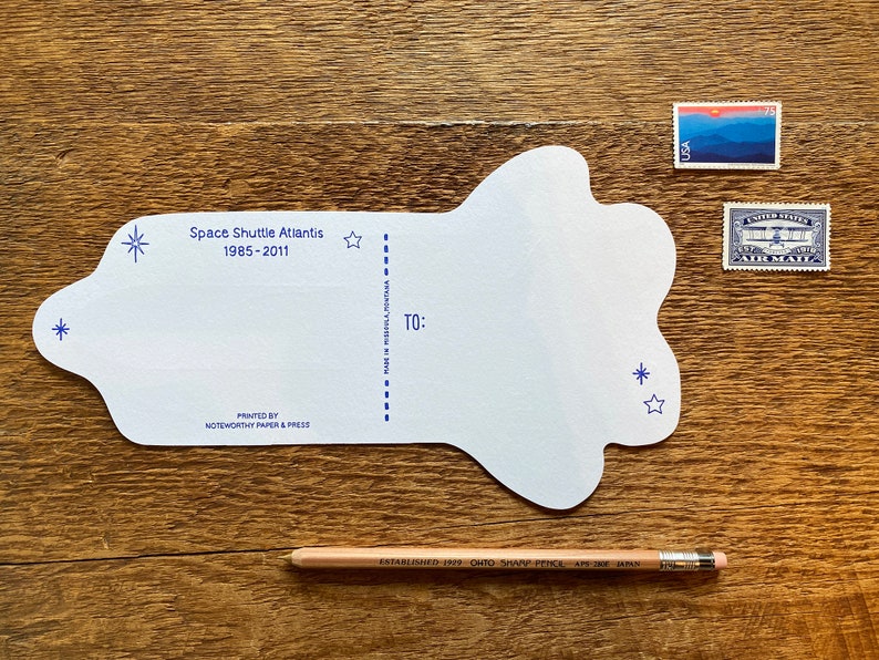 May include: A white postcard shaped like a cloud with blue stars and the text "Space Shuttle Atlantis 1985-2011" printed on it. The postcard has a space for a message and an address.
