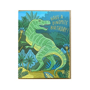 Op de afbeelding: Een groene dinosaurus met een tandenrij staat in een jungle-omgeving met palmbomen en blauwe bloemen. De tekst "Have a Dinomite Birthday!" is geschreven in gouden letters.
