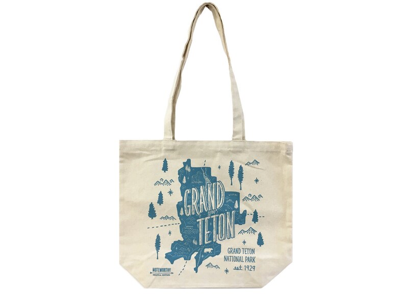 K&ouml;nnte beinhalten: Eine wei&szlig;e Canvas-Tragetasche mit einer blauen Grafik des Grand Teton National Park, Wyoming. Die Grafik enth&auml;lt den Text "Grand Teton" und das Gr&uuml;ndungsdatum des Parks, "est. 1929".