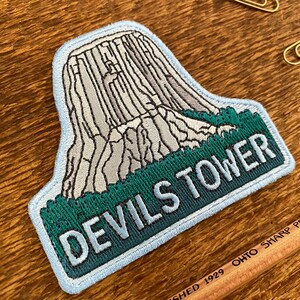 May include: A blue and green embroidered patch with a white outline. The patch features a depiction of Devils Tower National Monument, a large rock formation in Wyoming, with the text "Devils Tower" below.