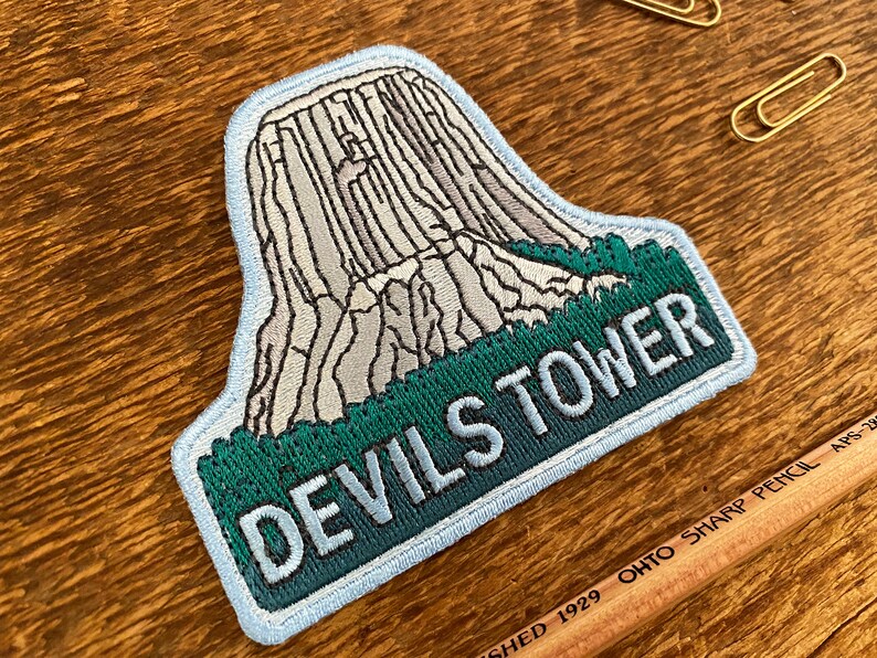 May include: A blue and green embroidered patch with a white outline. The patch features a depiction of Devils Tower National Monument, a large rock formation in Wyoming, with the text "Devils Tower" below.