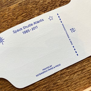 May include: A white card with blue text and stars. The text reads "Space Shuttle Atlantis 1985-2011" and "Printed by Noteworthy Paper & Press" and "Made in Missoula, Montana". The card has a dotted line and the word "TO:" printed on it.