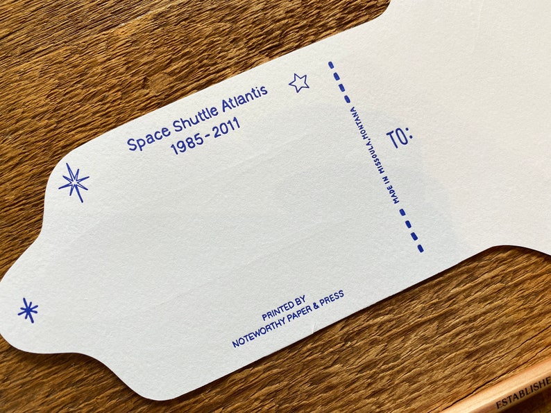 May include: A white card with blue text and stars. The text reads "Space Shuttle Atlantis 1985-2011" and "Printed by Noteworthy Paper & Press" and "Made in Missoula, Montana". The card has a dotted line and the word "TO:" printed on it.