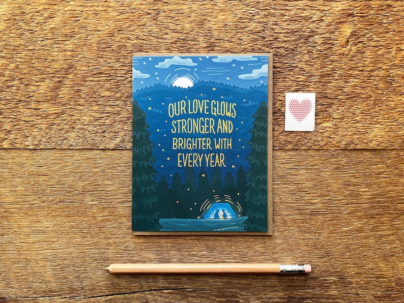 May include: A greeting card featuring a blue and green illustration of a starry night sky with a campfire and a tent. The text on the card reads "Our love glows stronger and brighter with every year."