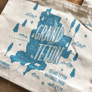 K&ouml;nnte beinhalten: Eine wei&szlig;e Canvas-Tragetasche mit einem blauen Grafikdesign des Grand Teton Nationalparks. Die Grafik enth&auml;lt eine Karte des Parks mit Bergen, B&auml;umen und dem Text "Grand Teton" und "est. 1929".