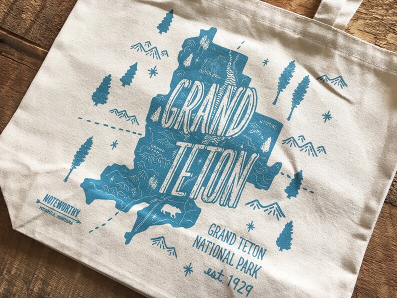 K&ouml;nnte beinhalten: Eine wei&szlig;e Canvas-Tragetasche mit einem blauen Grafikdesign des Grand Teton Nationalparks. Die Grafik enth&auml;lt eine Karte des Parks mit Bergen, B&auml;umen und dem Text "Grand Teton" und "est. 1929".