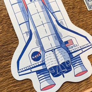 May include: A white and blue illustration of a space shuttle with the words "NASA" and "Atlantis" on it. The shuttle has red accents and is set against a woodgrain background.