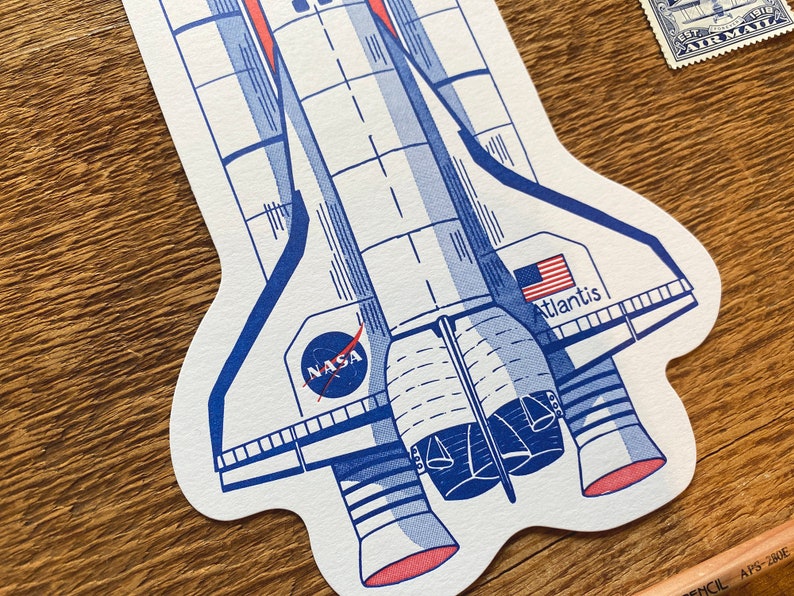 May include: A white and blue illustration of a space shuttle with the words "NASA" and "Atlantis" on it. The shuttle has red accents and is set against a woodgrain background.