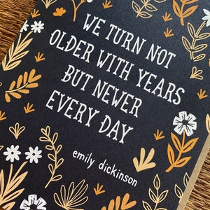 May include: A black card with a white floral pattern and the text "We turn not older with years but newer every day" by Emily Dickinson.