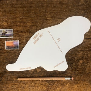 Peut inclure: Une carte blanche en forme de dinosaure avec le texte "Having a Dinomite Time!" et "To:" avec une ligne pointill&eacute;e pour &eacute;crire un nom. La carte est sur une surface en bois avec deux timbres-poste et un crayon.
