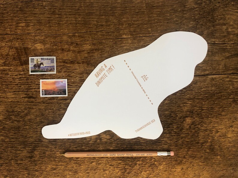 Peut inclure: Une carte blanche en forme de dinosaure avec le texte "Having a Dinomite Time!" et "To:" avec une ligne pointill&eacute;e pour &eacute;crire un nom. La carte est sur une surface en bois avec deux timbres-poste et un crayon.