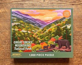 May include: A 1,000 piece jigsaw puzzle featuring a scenic view of the Great Smoky Mountains National Park. The puzzle depicts a colorful landscape with lush green trees, a winding road, and a sunset sky. Three black bears are visible in the foreground.