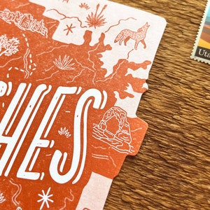 May include: A red and white postcard with a map of Arches National Park in Utah. The postcard features a stylized illustration of a coyote, a cactus, and a rock formation. The text "Arches" is written in large white letters across the top of the postcard. The postcard also features a stamp with a picture of Delicate Arch and the text "Utah 1896".