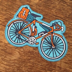 May include: A blue and orange embroidered patch of a bicycle with a backpack and sleeping bag attached to the back.
