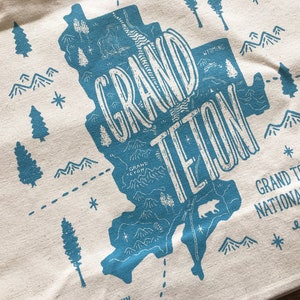 K&ouml;nnte beinhalten: Eine blaue und wei&szlig;e Tragetasche mit einer Karte des Grand Teton Nationalparks, Wyoming. Der Text "GRAND TETON" ist in einer gro&szlig;en, stilisierten Schrift geschrieben. Der Text "GRAND TETON NATIONAL PARK est. 1929" steht unter der Karte.