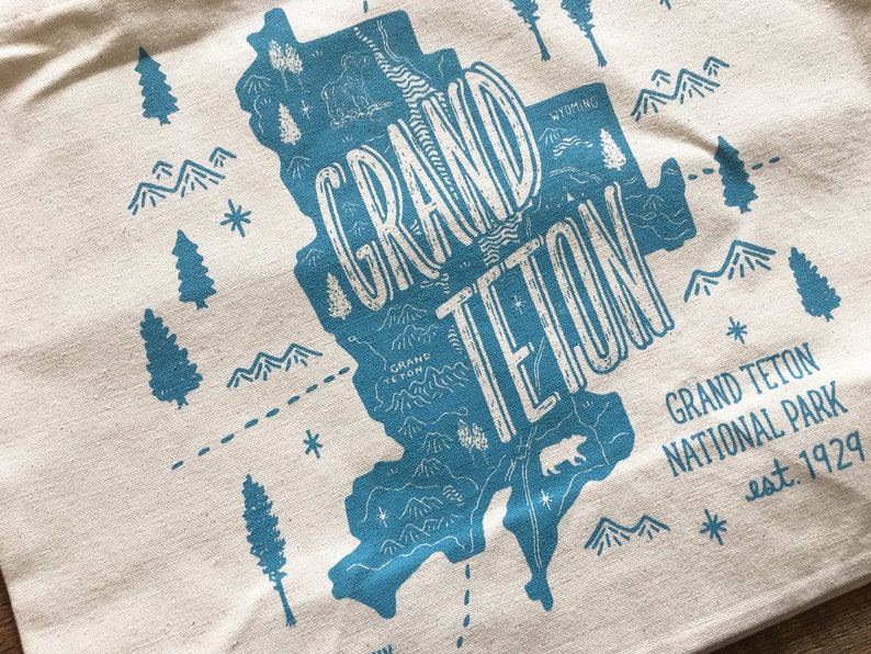 K&ouml;nnte beinhalten: Eine blaue und wei&szlig;e Tragetasche mit einer Karte des Grand Teton Nationalparks, Wyoming. Der Text "GRAND TETON" ist in einer gro&szlig;en, stilisierten Schrift geschrieben. Der Text "GRAND TETON NATIONAL PARK est. 1929" steht unter der Karte.