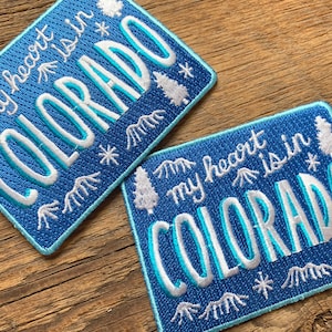 Può includere: Due toppe ricamate blu con lettere bianche. Le toppe presentano una catena montuosa, pini e fiocchi di neve. Il testo sulle toppe recita "My heart is in Colorado".