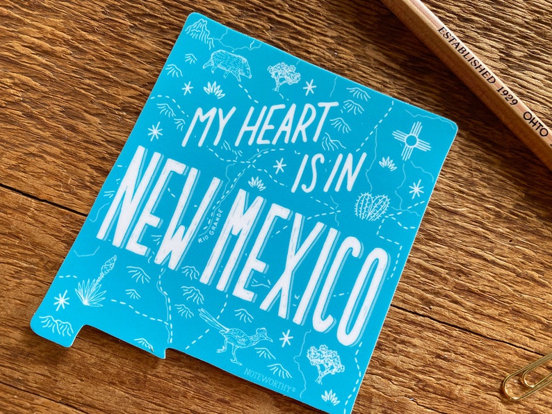 May include: A blue sticker with a white outline of the state of New Mexico. The sticker says "My heart is in New Mexico" with a stylized map of the state in the background.