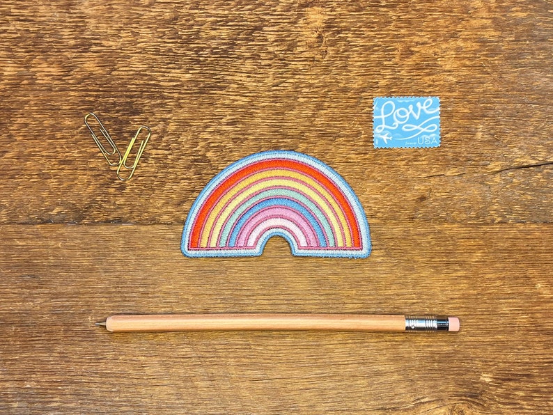Pu&ograve; includere: Una toppa ricamata a forma di arcobaleno con un contorno blu. La toppa &egrave; su una superficie di legno con una matita e due graffette. Un francobollo blu con la parola "Love" &egrave; nell'angolo in alto a destra.