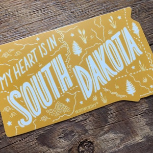 Pu&ograve; includere: Adesivo giallo con un contorno bianco dello stato del Dakota del Sud. L'adesivo dice "My heart is in South Dakota" e ha un piccolo logo "Noteworthy" in basso a destra.