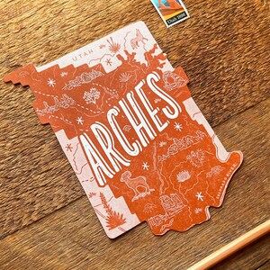 May include: A red and white illustrated map of Arches National Park in Utah. The map features a stylized outline of the park with various landmarks and details, including the Colorado River. The word "ARCHES" is written in large, stylized letters across the map.