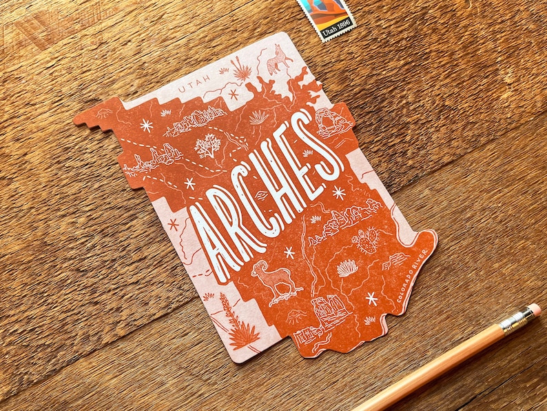 May include: A red and white illustrated map of Arches National Park in Utah. The map features a stylized outline of the park with various landmarks and details, including the Colorado River. The word "ARCHES" is written in large, stylized letters across the map.