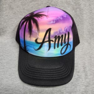 Peut inclure: Une casquette trucker noire avec un panneau avant orné d'un coucher de soleil à l'aérographe, une silhouette de palmier et le prénom Amy écrit en lettres cursives noires. La casquette possède une visière noire et des panneaux en filet.