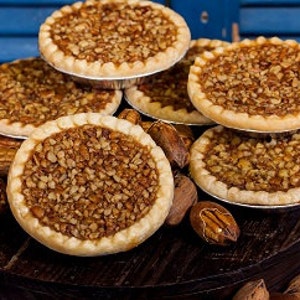 May include: A close-up of a stack of mini pecan pies on a wooden surface. The pies have a golden brown crust and are filled with a rich, nutty pecan filling. Pecan nuts are scattered around the pies.