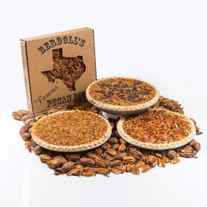 May include: Three pecan pies in individual pie tins on a white surface surrounded by pecan halves. The pies are topped with pecans and a brown sugary glaze. A cardboard box with the text "Berddoll's "Famous" Pecan Pie" is in the background.