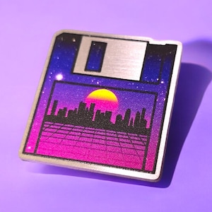 Puede incluir: Pin de disquete plateado con un diseño de paisaje urbano retro. El diseño presenta una puesta de sol con un sol amarillo, un horizonte de la ciudad y un patrón de cuadrícula, sobre un degradado de morado y rosa.