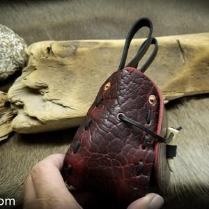 May include: A dark red leather pouch with black stitching and a flap closure. The pouch has a textured surface and is held in a hand.