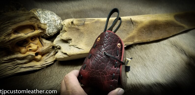 May include: A dark red leather pouch with black stitching and a flap closure. The pouch has a textured surface and is held in a hand.