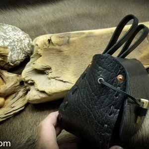 May include: A black leather pouch with a flap closure and two leather straps. The pouch has a decorative stitching pattern and copper rivets.