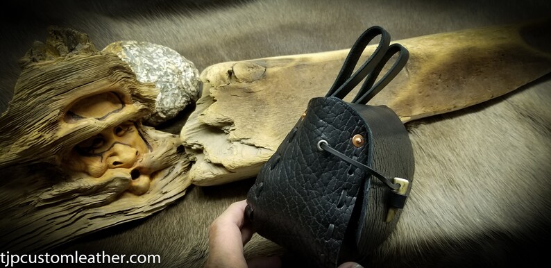 May include: A black leather pouch with a flap closure and two leather straps. The pouch has a decorative stitching pattern and copper rivets.