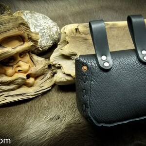 May include: Black leather pouch with two straps and silver rivets. The pouch is closed with a flap and has a decorative stitch around the edge.