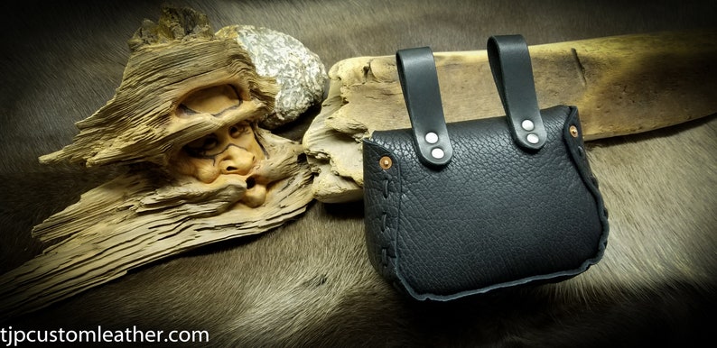 May include: Black leather pouch with two straps and silver rivets. The pouch is closed with a flap and has a decorative stitch around the edge.