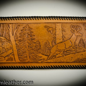 May include: A brown leather wallet with a tooled design of a deer in a woodland setting. The wallet has a black stitched border.