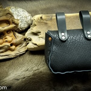 May include: Black leather pouch with two straps and silver rivets. The pouch is closed and has a decorative stitching around the edges.