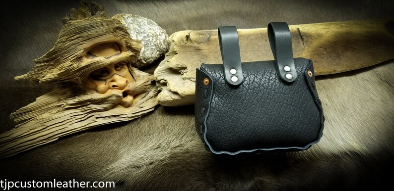 May include: Black leather pouch with two straps and silver rivets. The pouch is closed and has a decorative stitching around the edges.