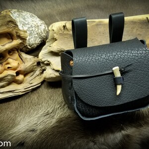 May include: A black leather pouch with a bone toggle closure. The pouch has two leather straps for attaching to a belt or other item.