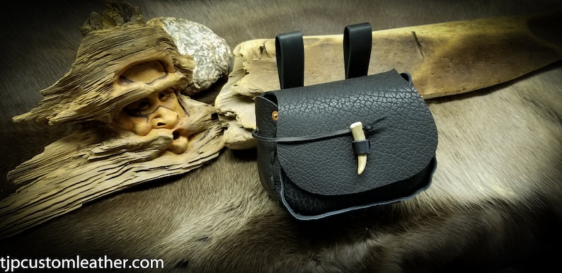 May include: A black leather pouch with a bone toggle closure. The pouch has two leather straps for attaching to a belt or other item.