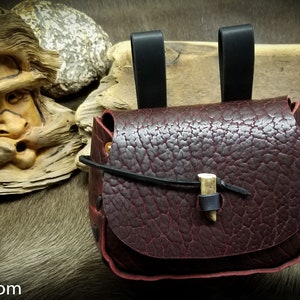 May include: A burgundy leather pouch with a bone toggle closure and two black leather straps. The pouch is sitting on a brown fur surface.