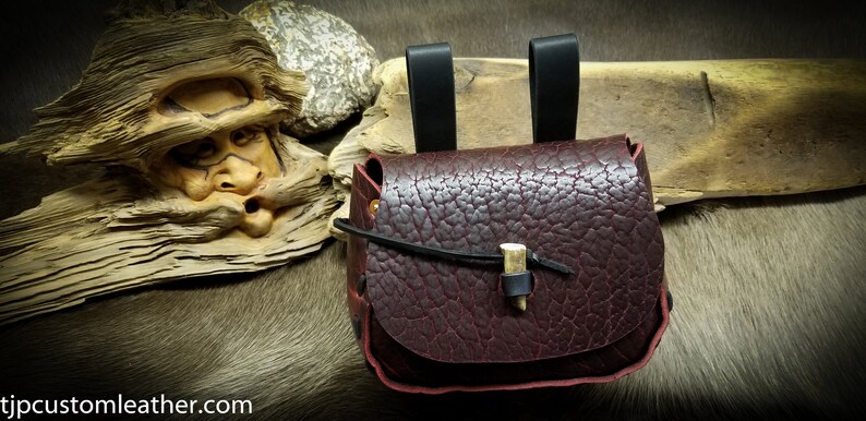 May include: A burgundy leather pouch with a bone toggle closure and two black leather straps. The pouch is sitting on a brown fur surface.