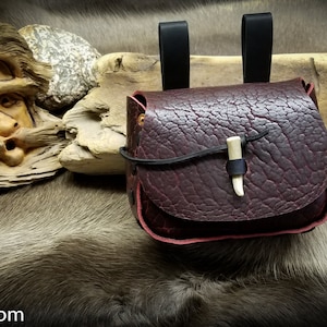 May include: A small, dark brown leather pouch with a flap closure and a bone toggle. The pouch has two black leather straps for carrying.