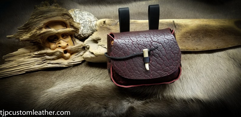 May include: A small, dark brown leather pouch with a flap closure and a bone toggle. The pouch has two black leather straps for carrying.