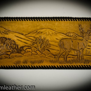 May include: A brown leather wallet with a tooled design of a deer in a mountain landscape. The wallet has a black stitched border.