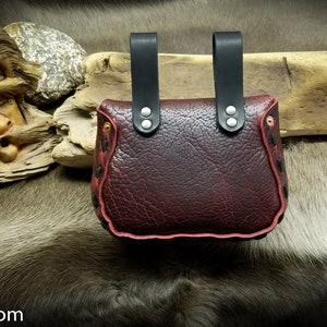 May include: A burgundy leather pouch with black leather straps and silver rivets. The pouch has a red stitched edge and is decorated with a pattern of small black dots.