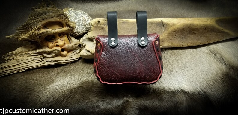 May include: A burgundy leather pouch with black leather straps and silver rivets. The pouch has a red stitched edge and is decorated with a pattern of small black dots.